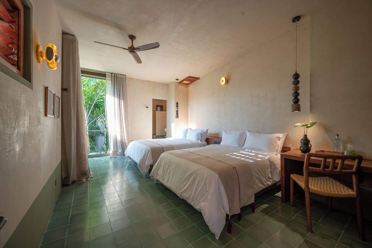 A hotel room with two beds, a ceiling fan, a wooden nightstand with a lamp, and a view of greenery outside.