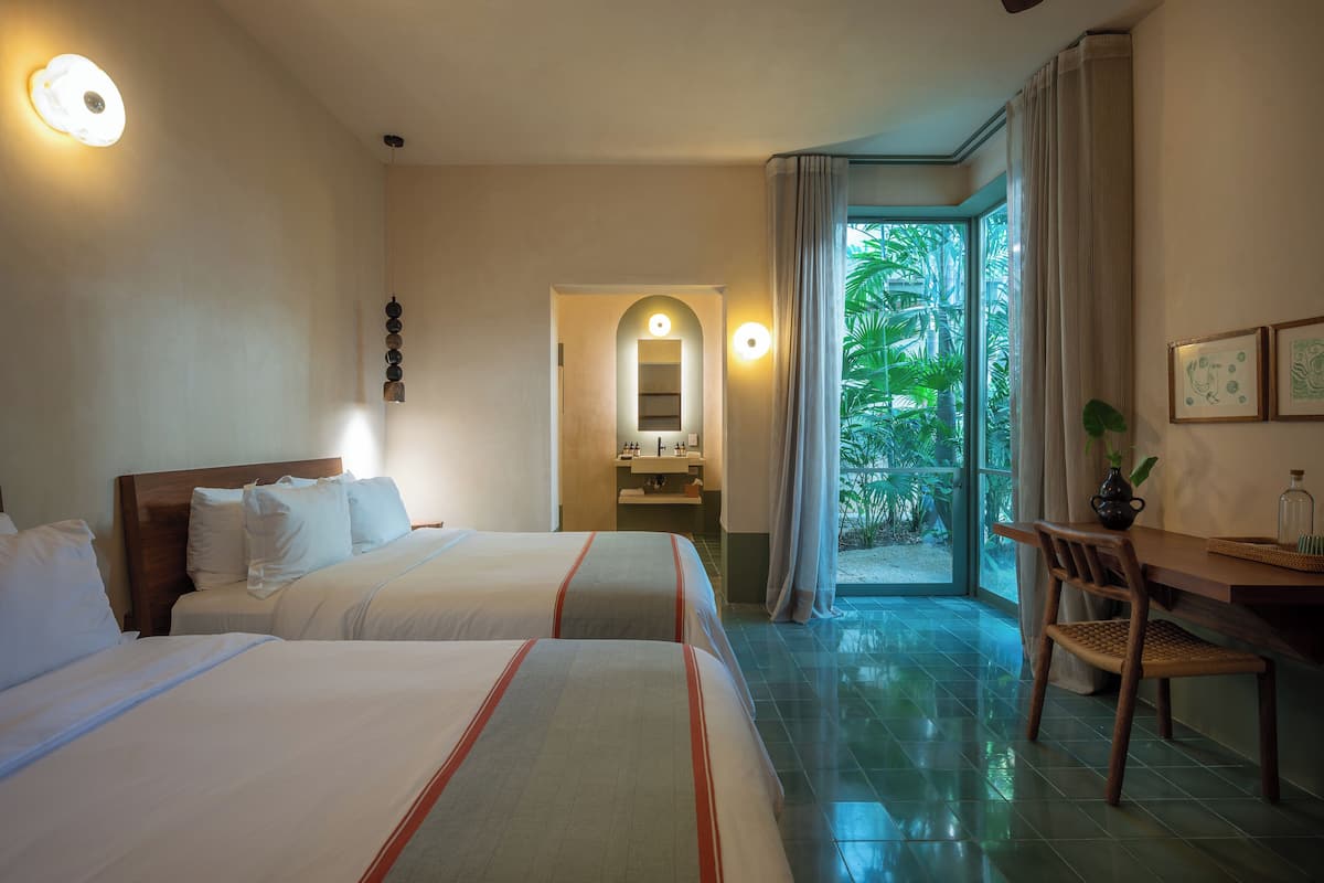 A hotel room with two beds, a desk, a chair, a bathroom, and a view of greenery.