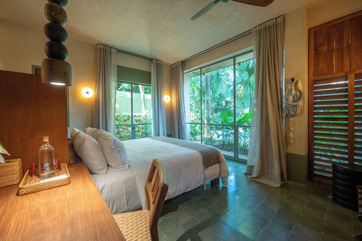 A bedroom with a bed, a wooden table with a bottle, a chair, and a window with curtains.