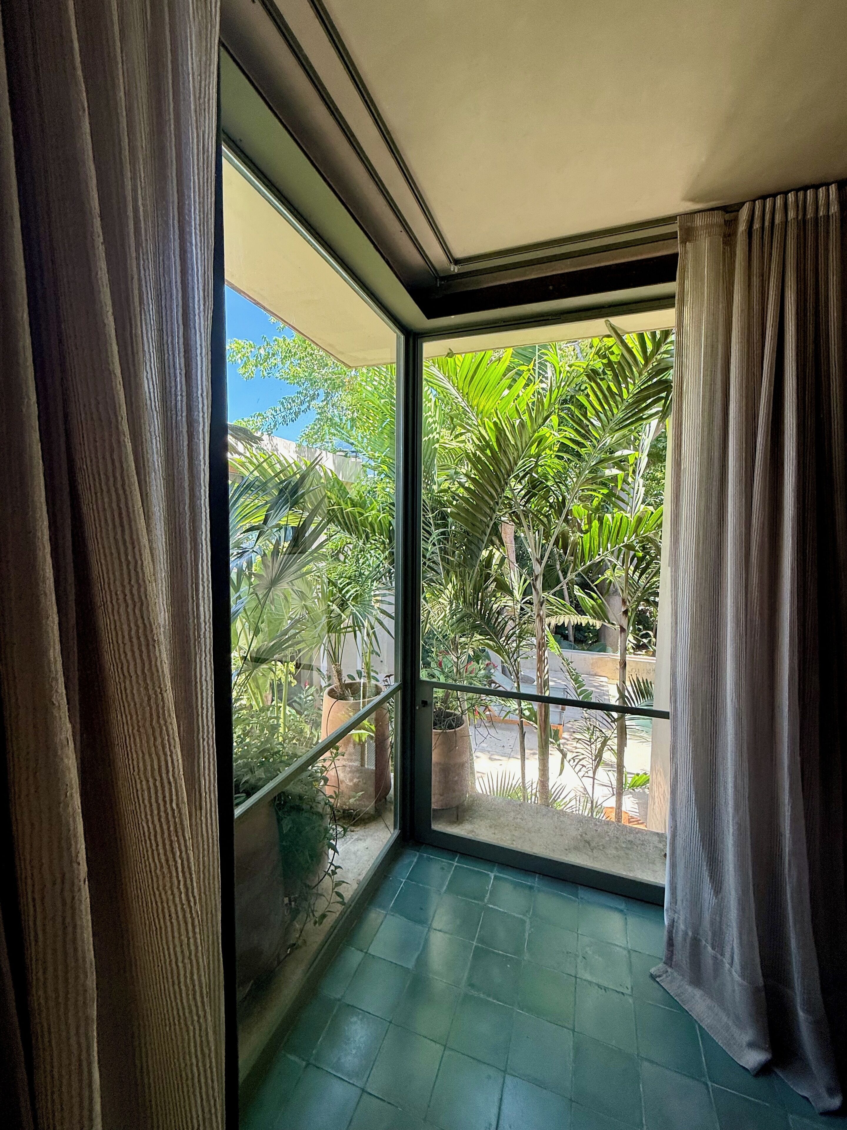 Una habitación con un gran ventanal que da a un balcón con plantas en macetas y vistas al verdor.