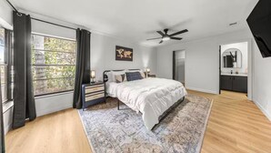 Une chambre à coucher comprenant un lit, des tables de chevet, un ventilateur de plafond, une fenêtre avec des rideaux et une vue sur l’extérieur.