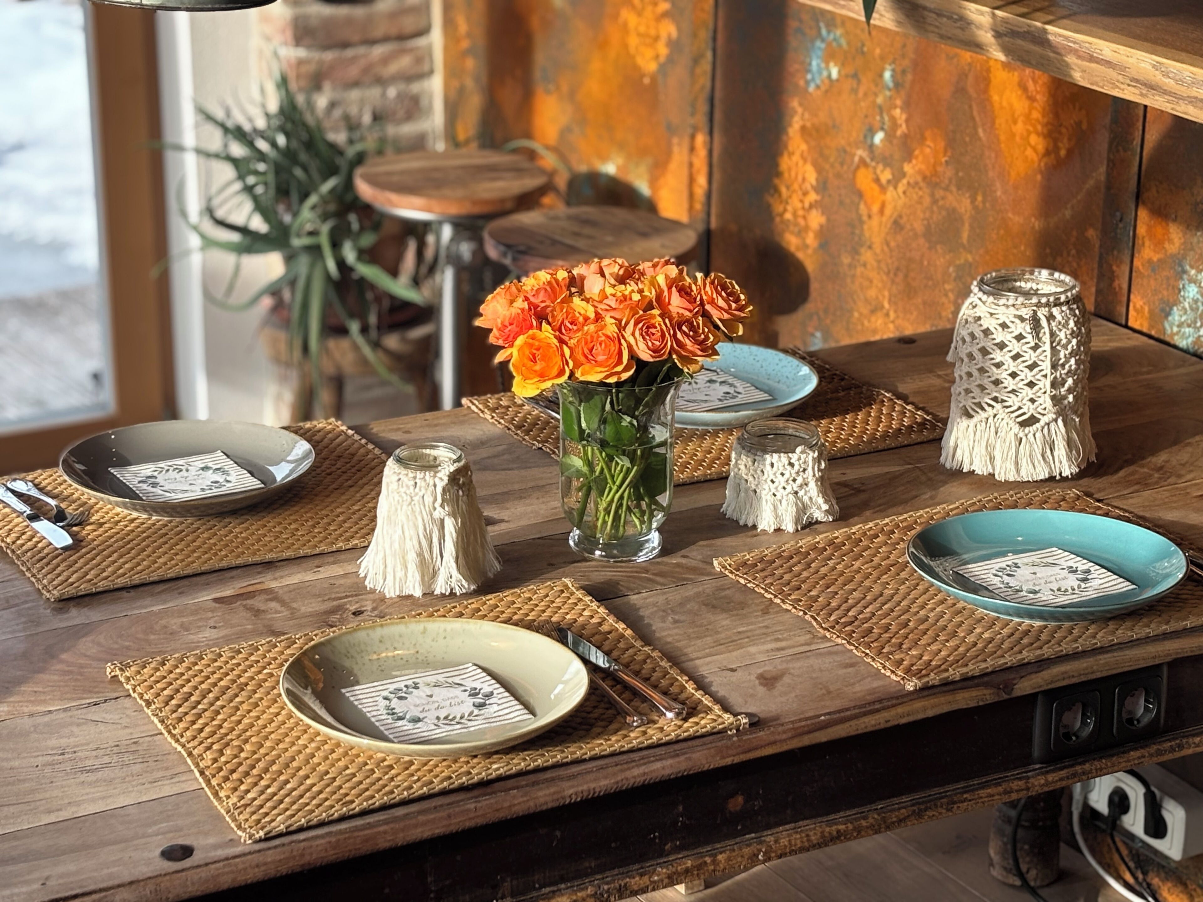 Une table à manger en bois, agrémentée de sets de table en fibres tressées, de vases en verre transparent ornés de fleurs oranges et d’assiettes en céramique aux motifs floraux.