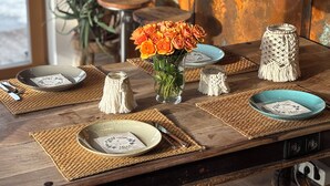 Une table à manger en bois, agrémentée de sets de table en fibres tressées, de vases en verre transparent ornés de fleurs oranges et d’assiettes en céramique aux motifs floraux.