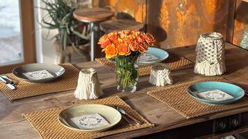 Une table à manger en bois, agrémentée de sets de table en fibres tressées, de vases en verre transparent ornés de fleurs oranges et d’assiettes en céramique aux motifs floraux.