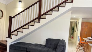 A spacious living area with a large gray sectional sofa, a wooden staircase with white balusters, and a dining area with orange chairs. - Luxury Retreat at Sorrell Ave (St Ann’s Bay)