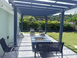 Ein überdachter Patio-Bereich mit Außenmöbeln, einer Feuerstelle und Blick auf einen üppigen Garten und Hügel im Hintergrund.