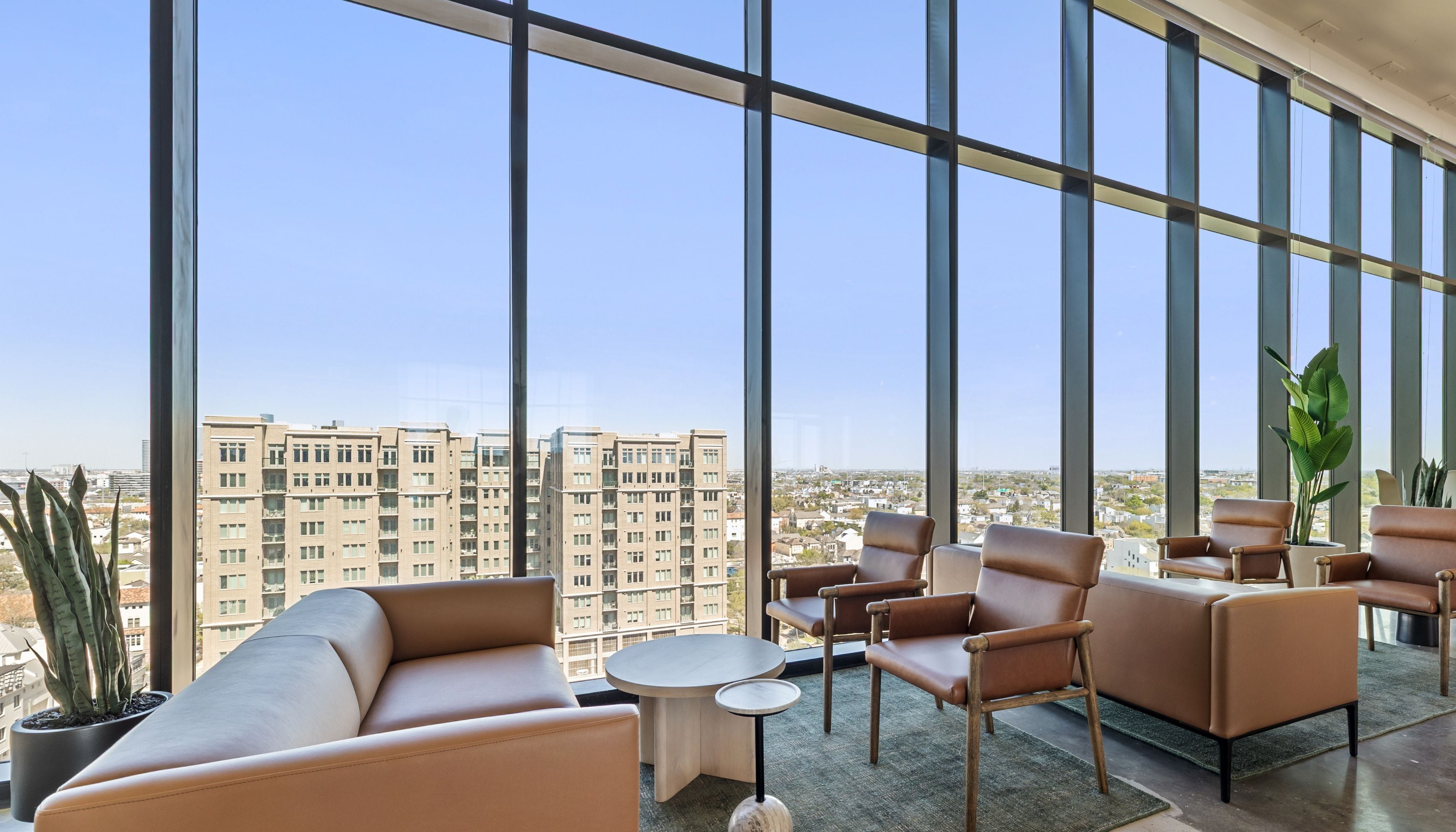 A high-rise hotel room with large windows, leather seating, a small round table, and a potted plant.