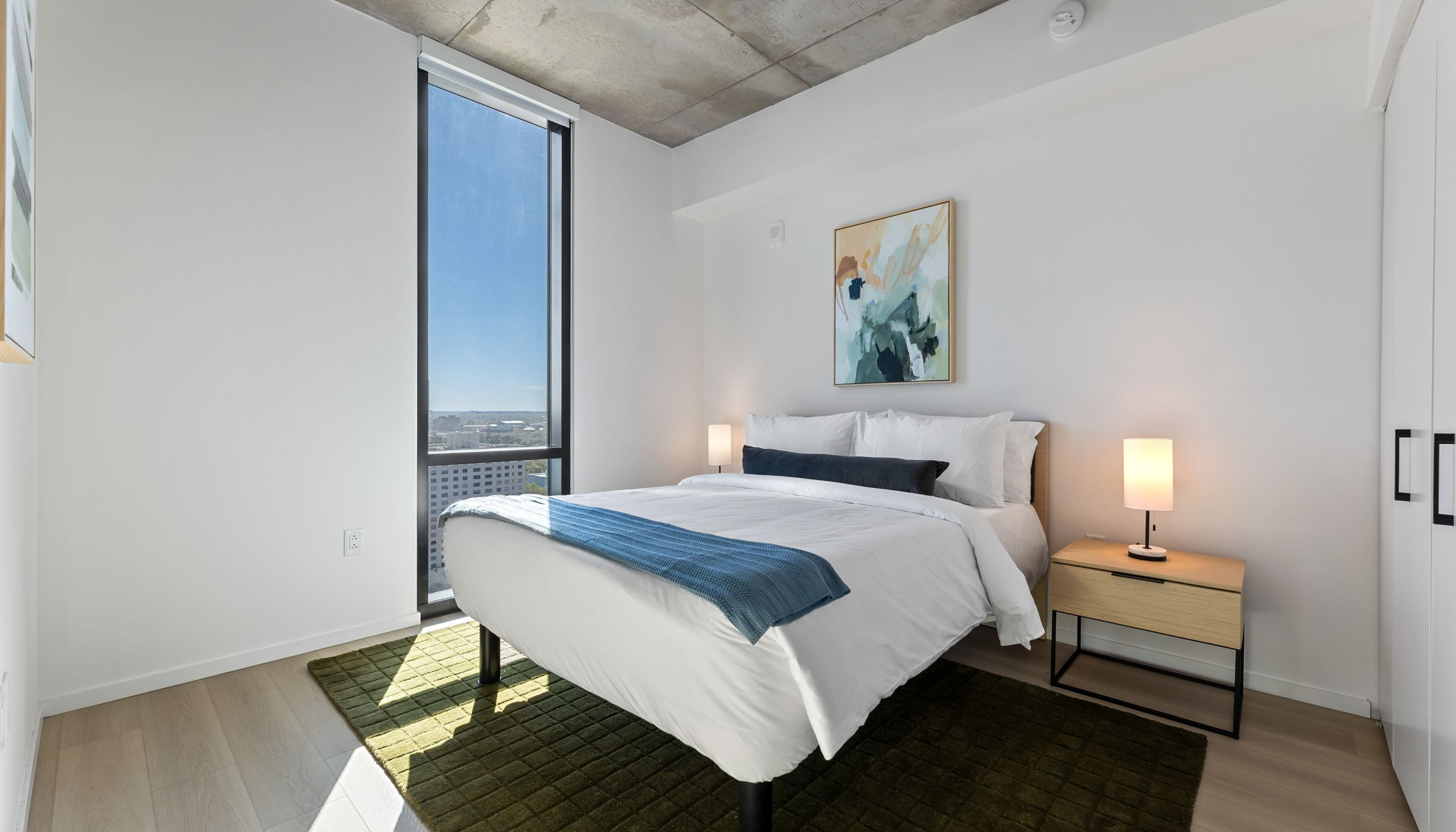 A modern hotel room with a large bed, a nightstand, a painting, and a view of the city.