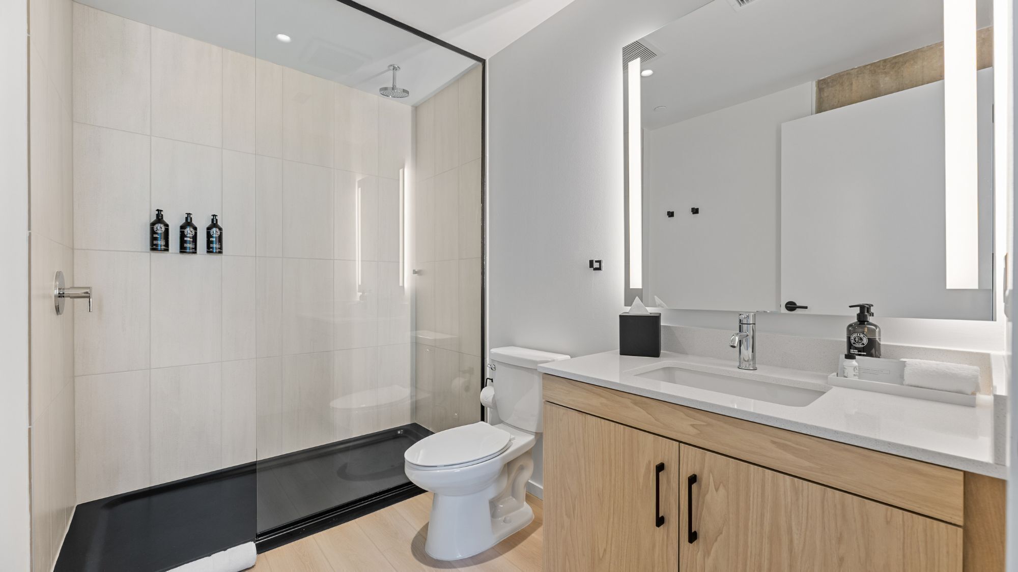 A modern bathroom with a glass shower enclosure, a white toilet, a vanity with a mirror, and toiletries on the counter.