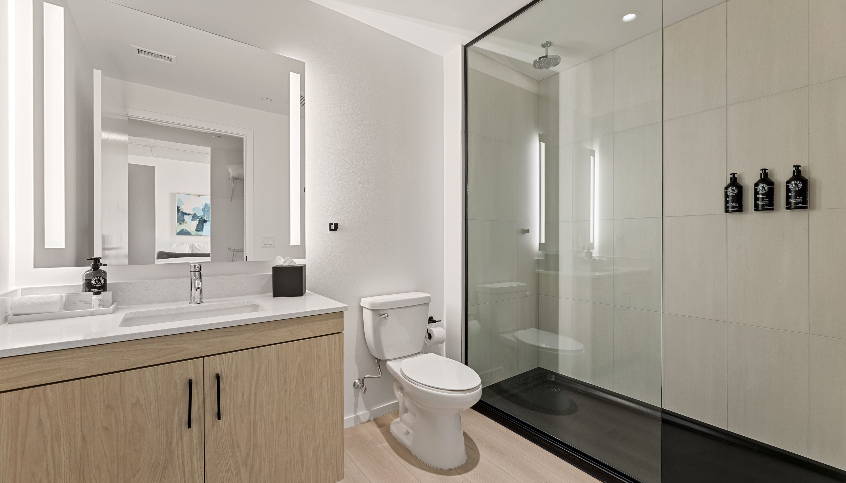 A modern bathroom with a white sink, mirror, and toilet, a glass shower enclosure, and three black bottles on the wall.