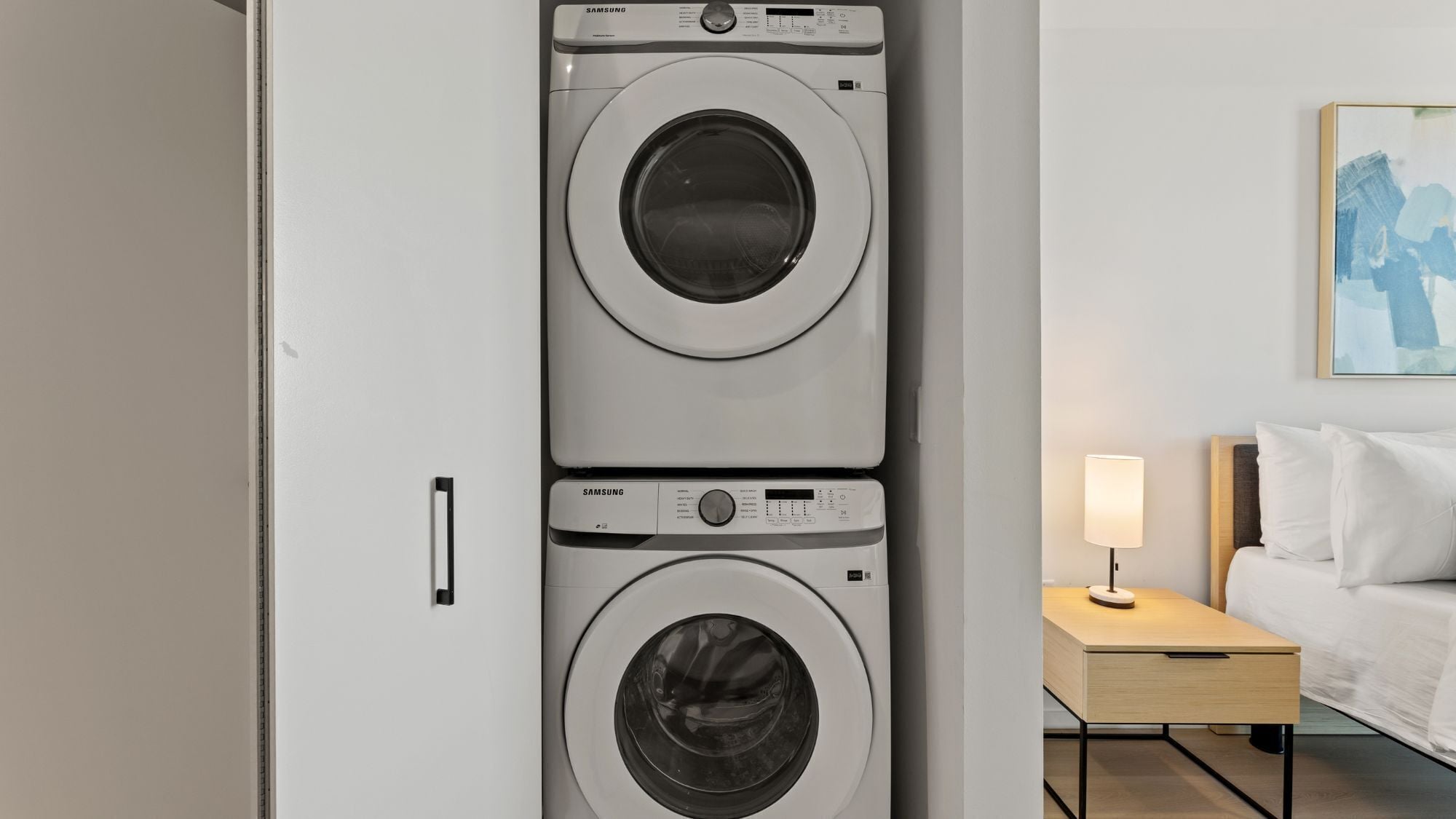 Une chambre d’hôtel équipée d’un lave-linge double, d’une table de chevet avec une lampe et d’un lit avec du linge de lit blanc.