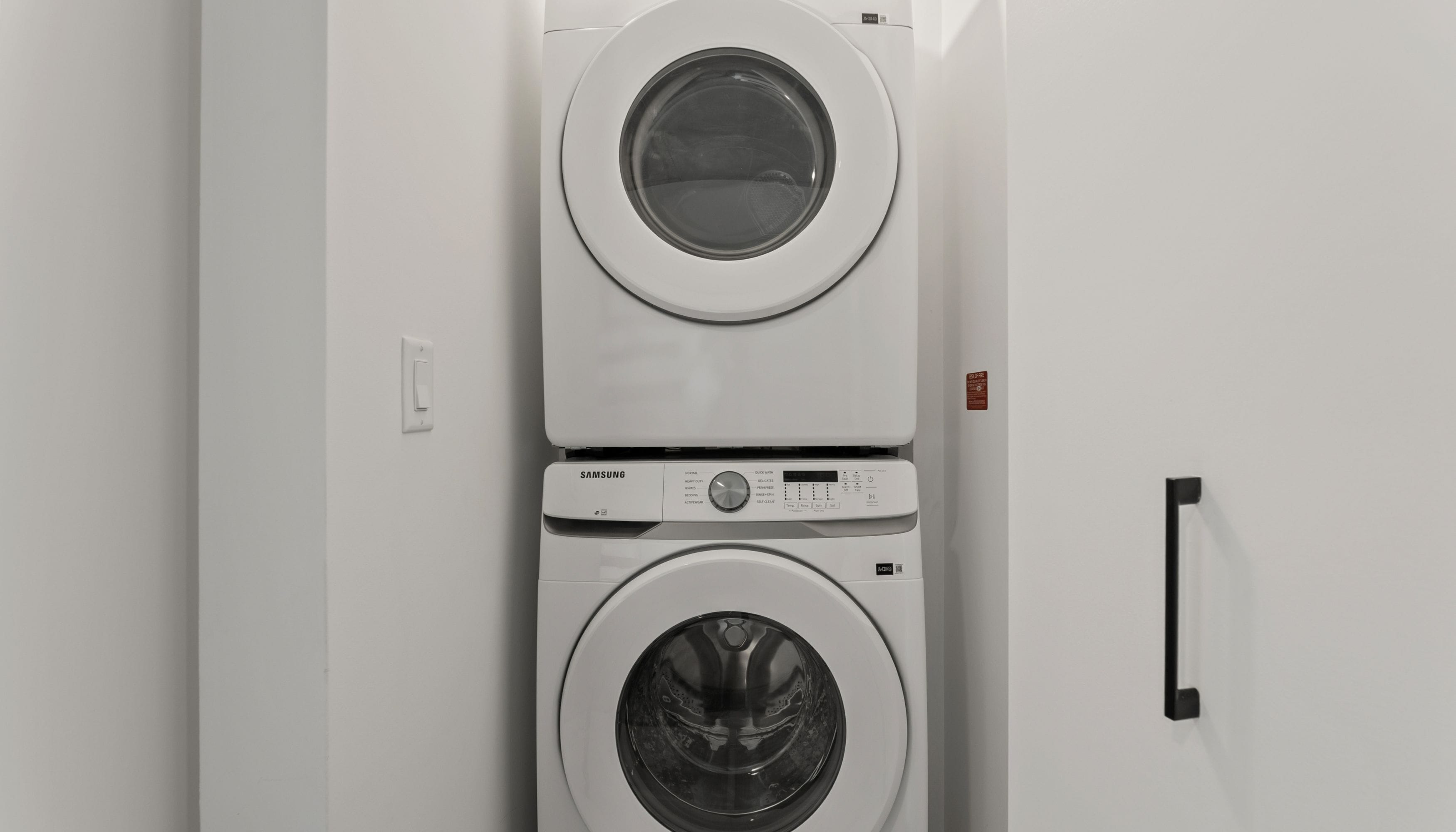 A front view of a stacked washer and dryer unit with a control panel, a power outlet, and a safety warning label.