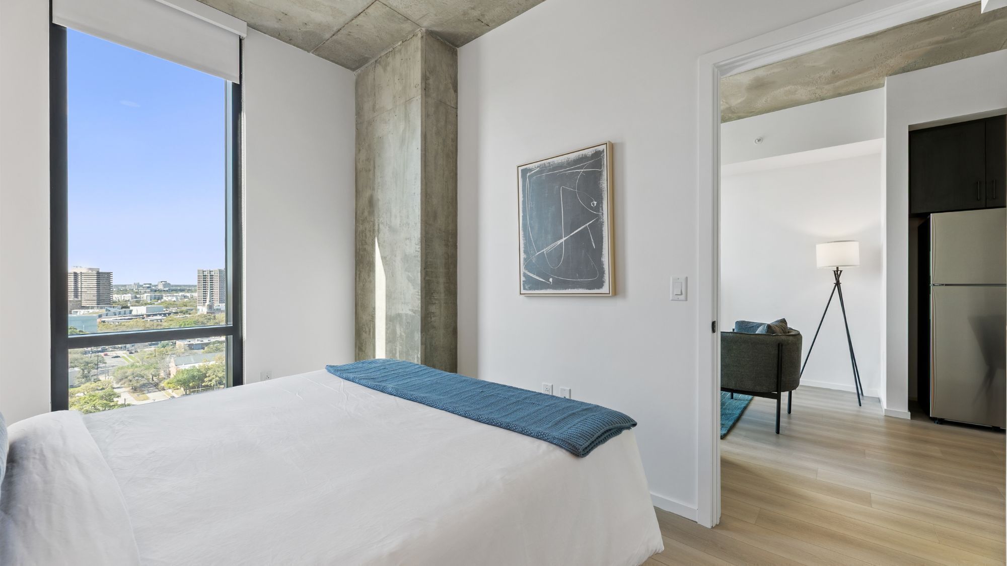 A modern hotel room with a large bed, a city view, a framed abstract painting, and a kitchenette.