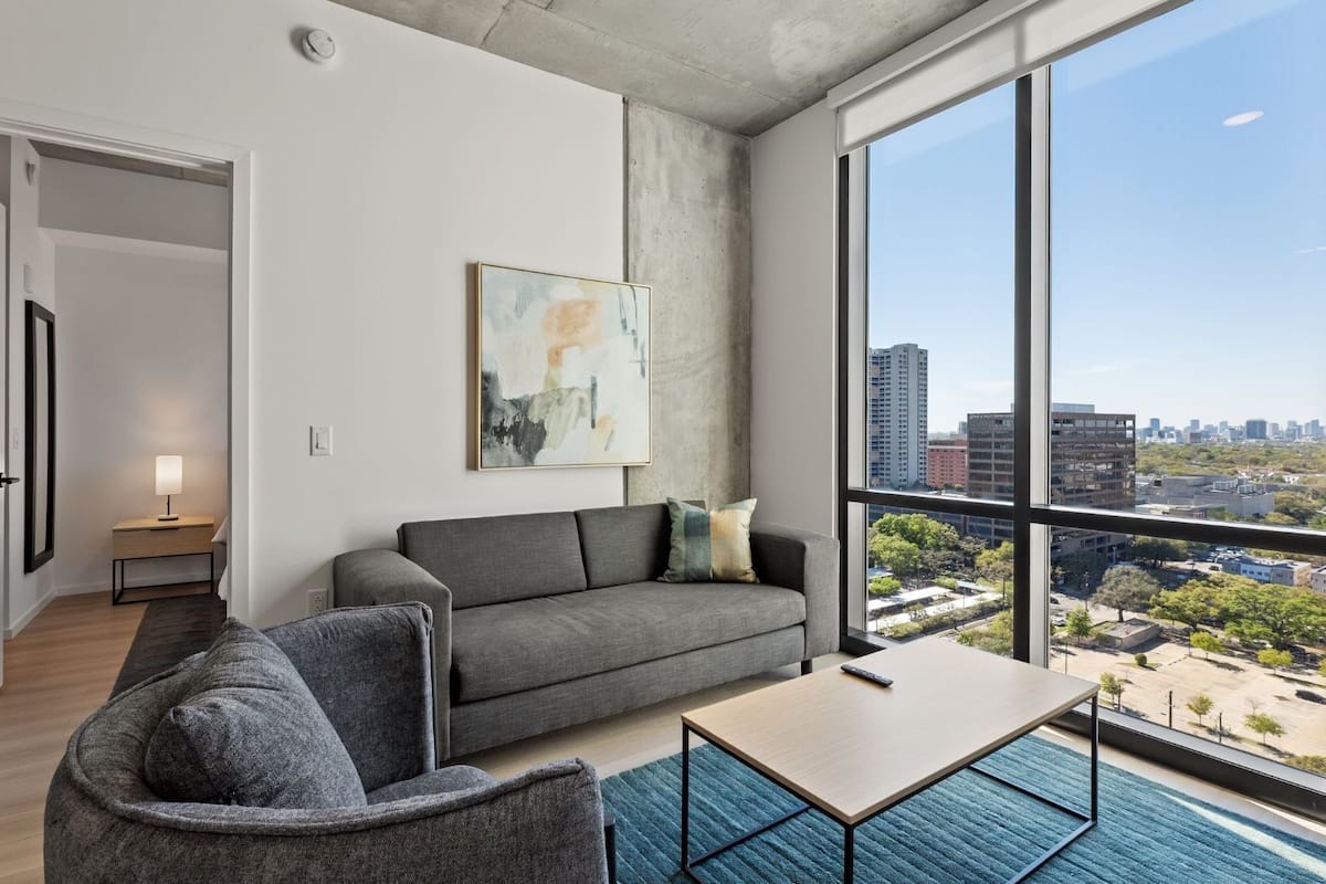 Una sala moderna con un sofá gris, una mesita y un ventanal con vistas a la ciudad.