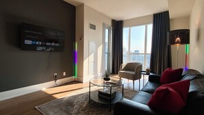 A modern living room with a flat-screen TV mounted on the wall, a sofa with red cushions, a coffee table, and a view of the city through large windows.