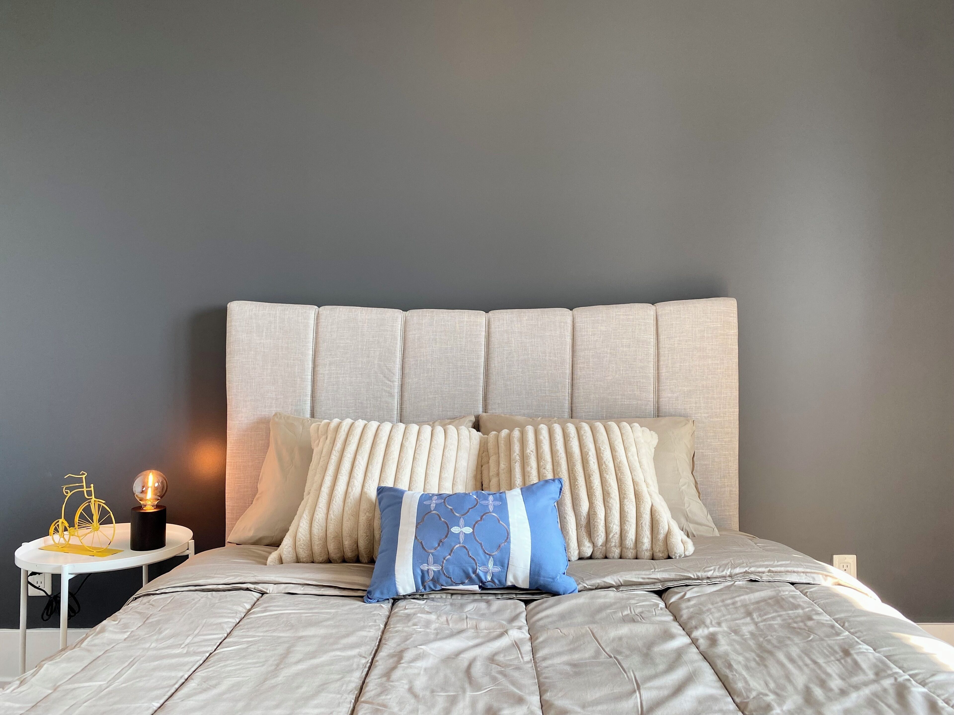 A neatly made bed with a grey headboard and multiple pillows, a small bedside table with a lamp, and a wall socket.
