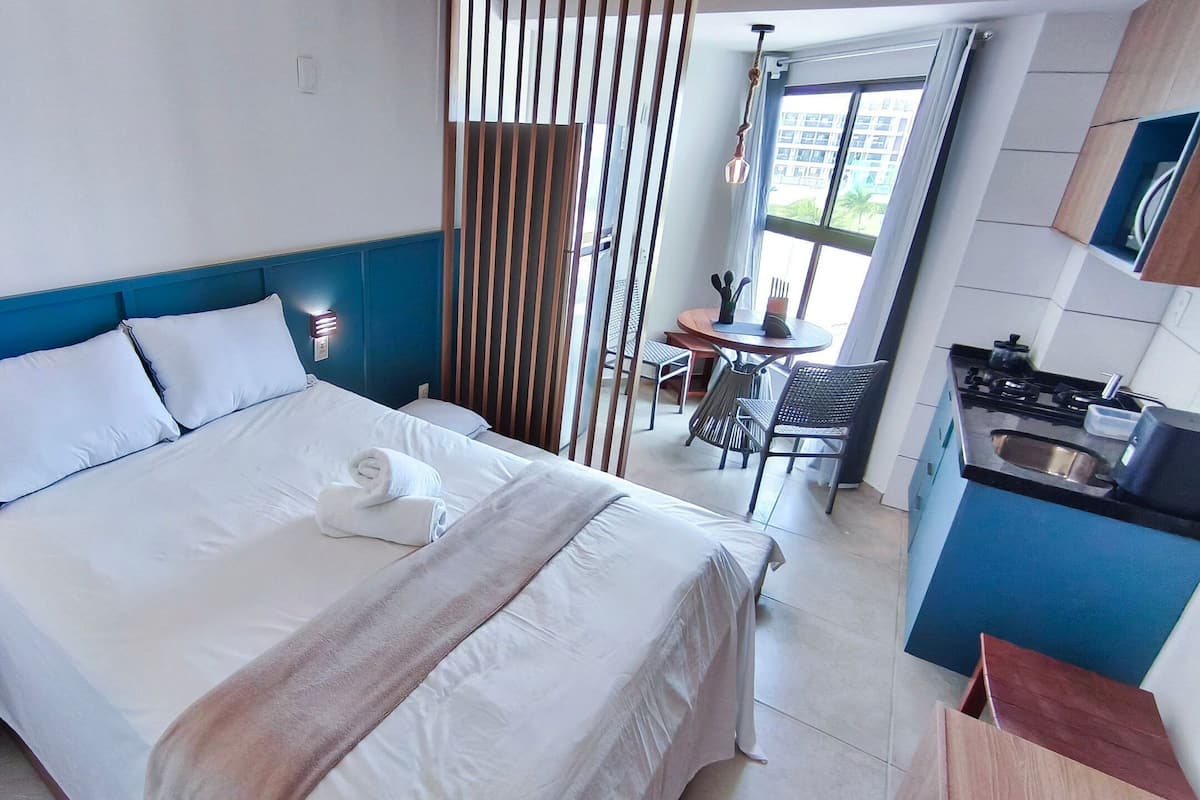 A hotel room with a bed, a small table, a chair, a kitchen area, and a window.