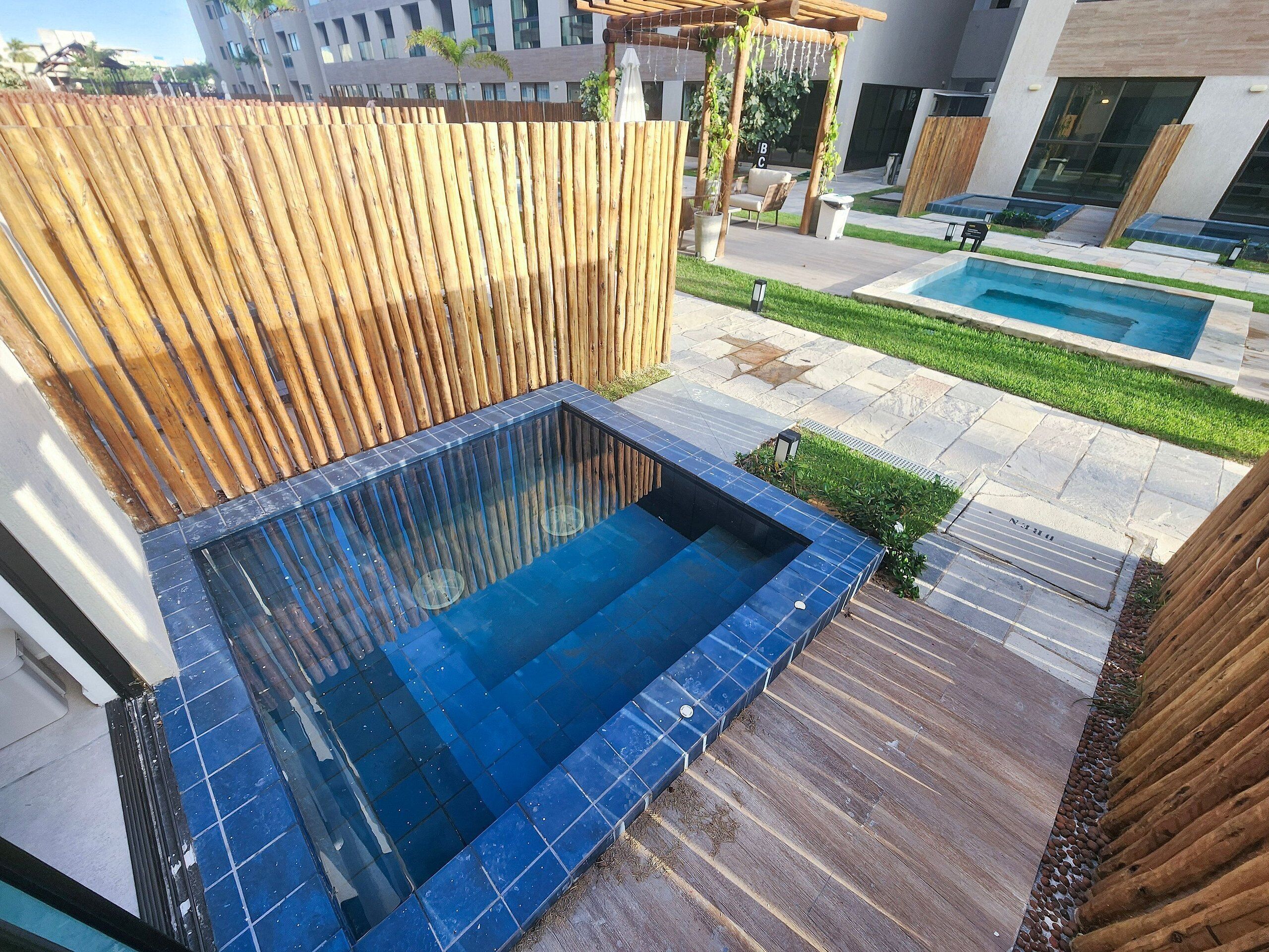 A rooftop pool area with a wooden pergola, a small seating area, and a view of a modern building in the background.