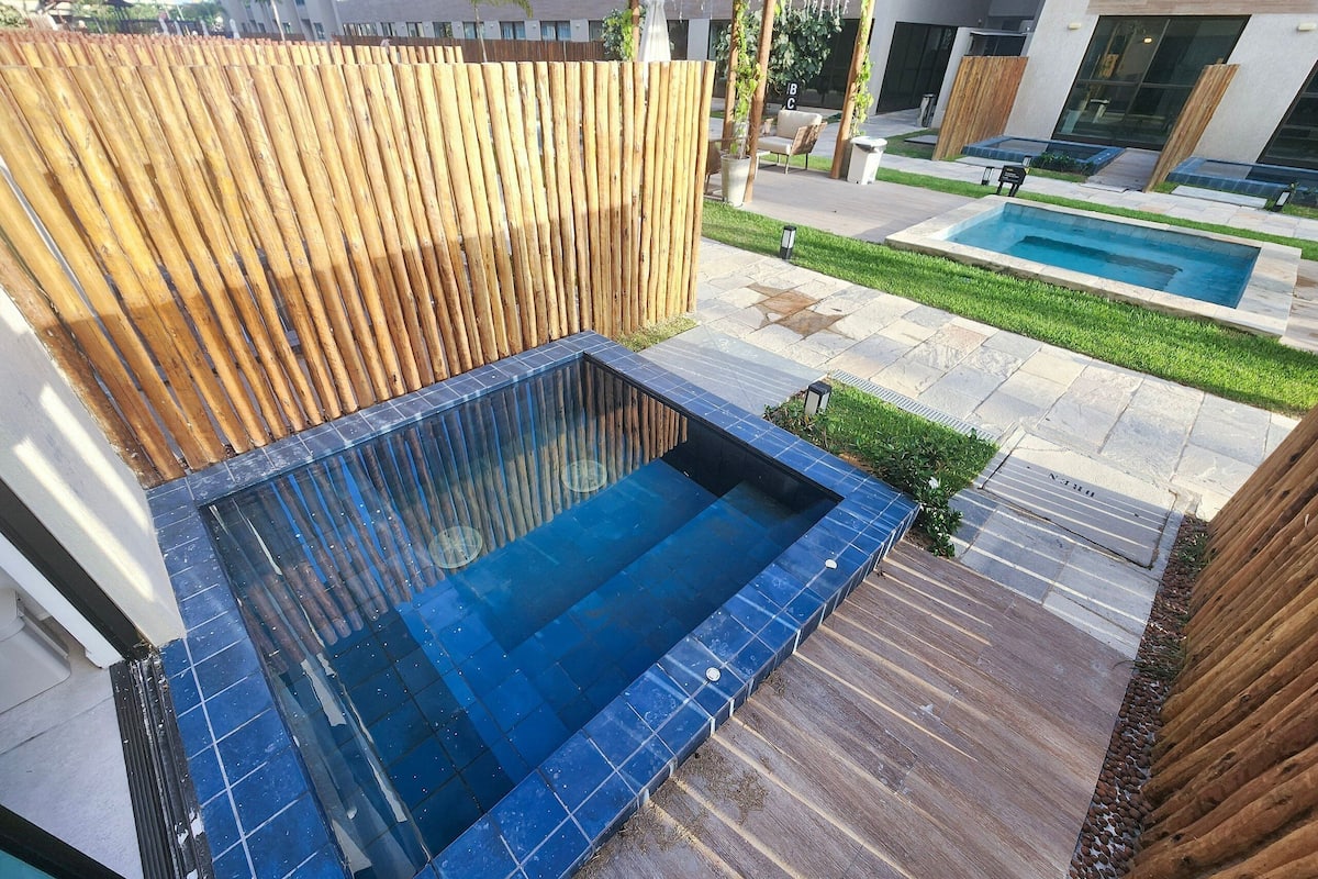 A rooftop pool area with a wooden pergola, a small seating area, and a view of a modern building in the background.