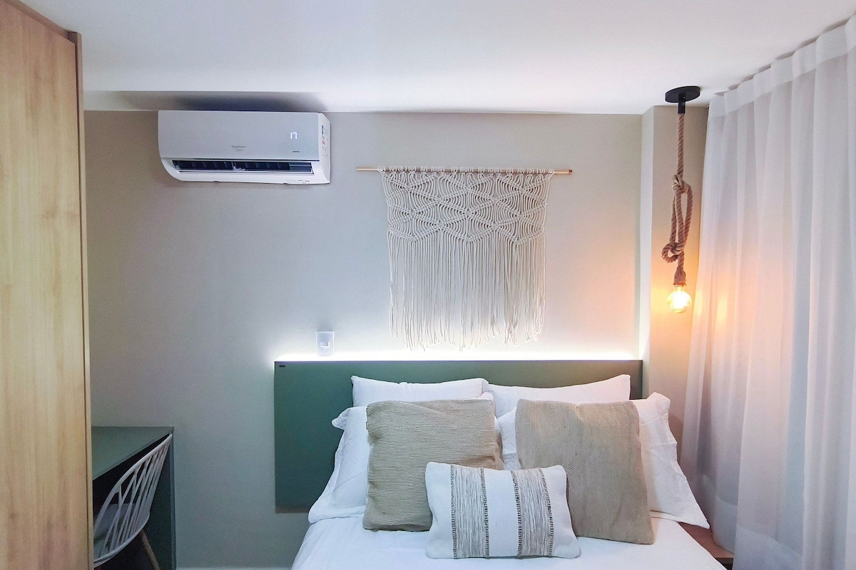 A hotel room with a bed, a wall-mounted air conditioner, a macrame wall hanging, and a hanging light fixture.