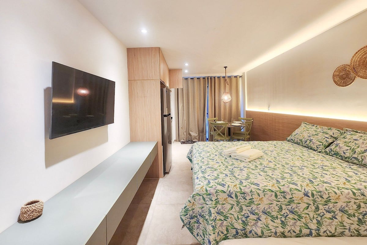 A hotel room with a bed, a TV mounted on the wall, a small table with chairs, and a kitchenette area.