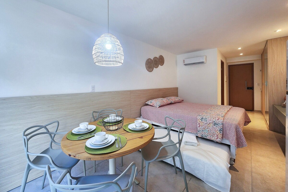 A compact room with a bed, a dining table with chairs, a hanging light fixture, and a door leading to another room.