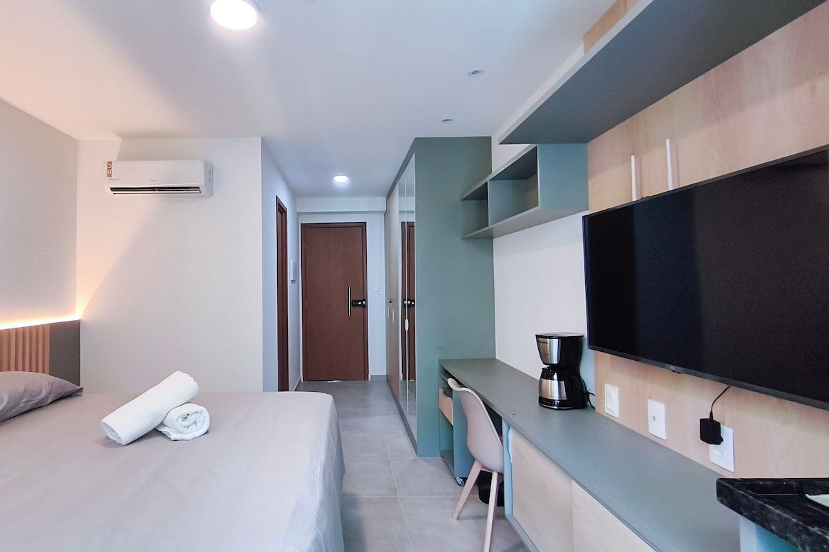 A hotel room with a bed, a desk, a chair, a flat-screen TV, and an air conditioning unit.