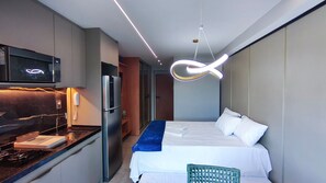 Kompakti hotellihuone, jossa on pieni keittiönurkkaus, valkoisilla lakanoilla varustettu sänky ja ainutlaatuinen kattovalaisin.