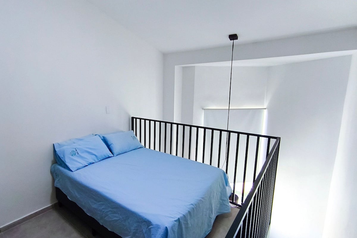 A small, single-bed room with a black metal headboard, a blue bedspread, and a window with a view.