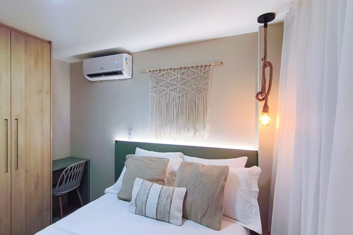 A bedroom with a bed, a desk, an air conditioner, and a macrame wall hanging.