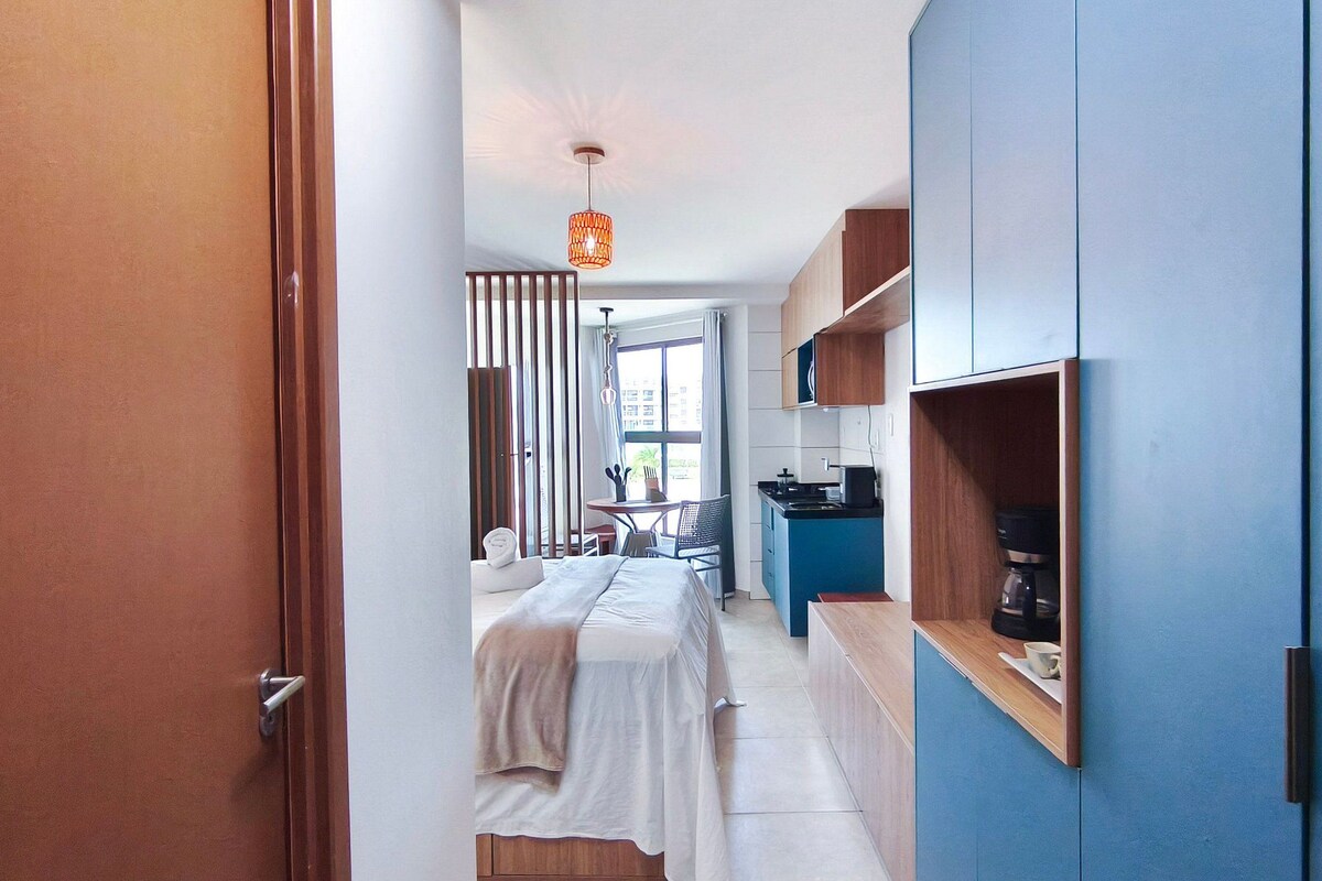 A narrow hallway leading to a bedroom with a bed, a small table, and a chair. There is a kitchenette with a stove and a coffee maker.