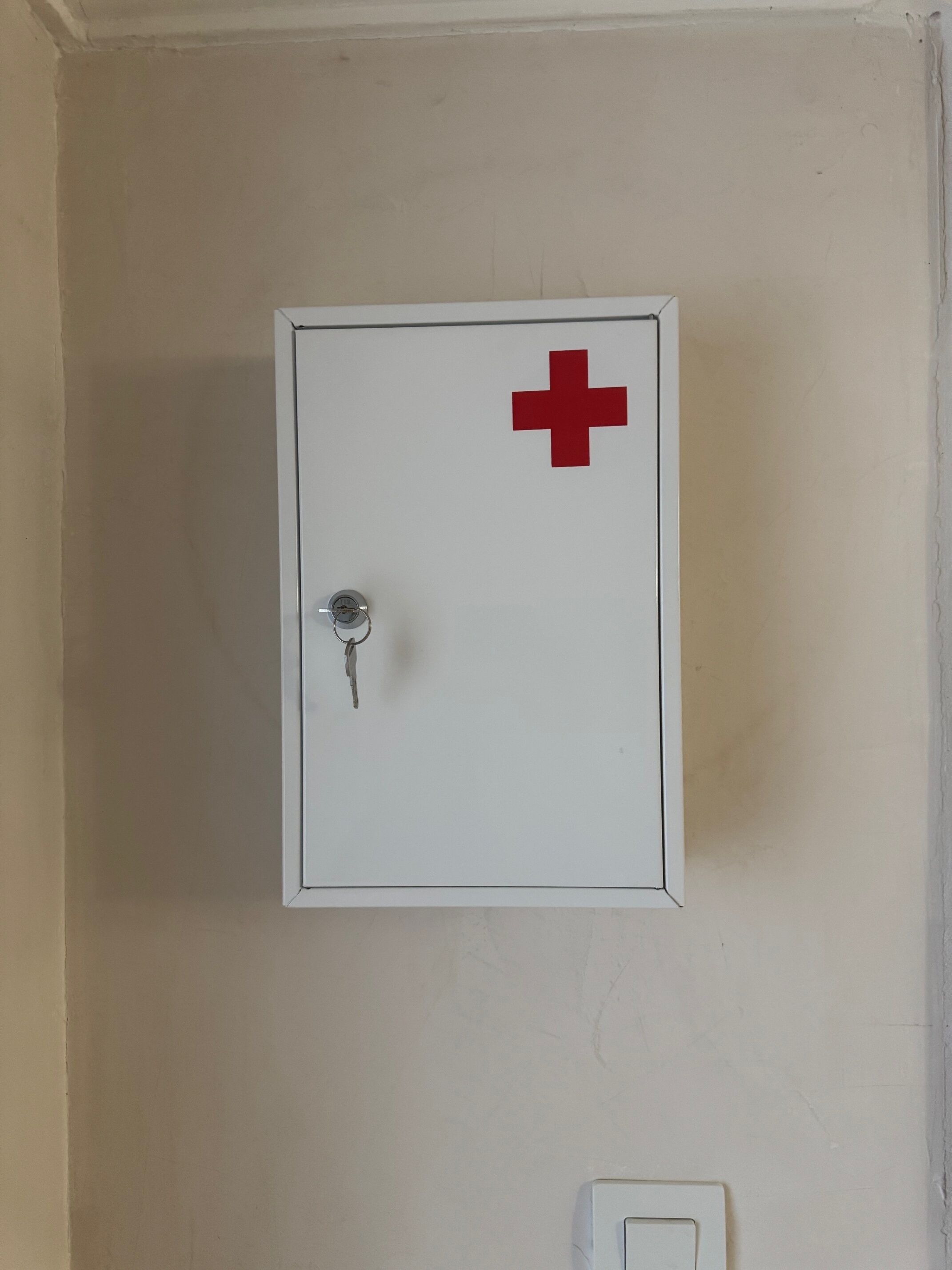 A white emergency medical kit with a red cross symbol, mounted on a beige wall.