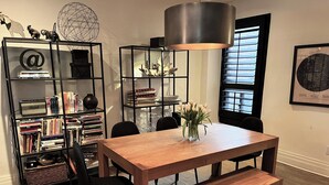 A dining area with a wooden table, black chairs, and a bench. There is a bookshelf filled with books and decorative items. A large lamp hangs above the table, and a window with blinds is visible in the background. - Trendy Roncesvalles Village. Just steps to shopping, dining, Lake Ontario (Toronto)