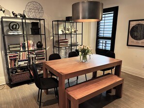 A dining area with a wooden table, black chairs, and a bench. There is a bookshelf filled with books and decorative items. A large lamp hangs above the table, and a window with blinds is visible in the background. - Trendy Roncesvalles Village. Just steps to shopping, dining, Lake Ontario (Toronto)
