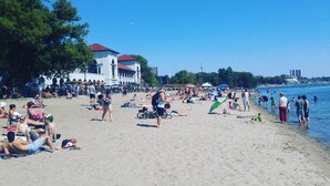 A beach with people sunbathing and swimming, a large building with a red roof, and a clear blue sky. - Trendy Roncesvalles Village. Just steps to shopping, dining, Lake Ontario (Toronto)