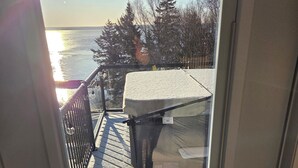A view from inside a room looking out to a balcony with a metal railing, a snow-covered roof, and a body of water in the background.