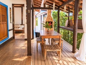 A wooden deck with a table and chairs, a brick fireplace, and a view of greenery. - House with a Heated Pool in Camburi | River and Nature (São Sebastião)
