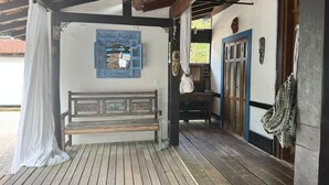 En träbänk under en överbyggd veranda, med en dekorativ spegel och en dörr med fönsterluckor.