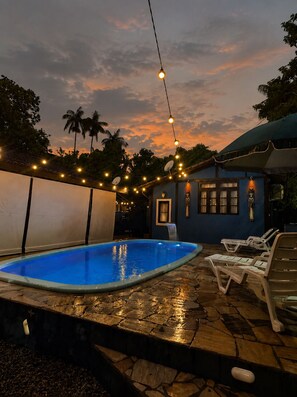 A swimming pool with lounge chairs, a stone deck, and string lights against a sunset sky. - House with a Heated Pool in Camburi | River and Nature (São Sebastião)