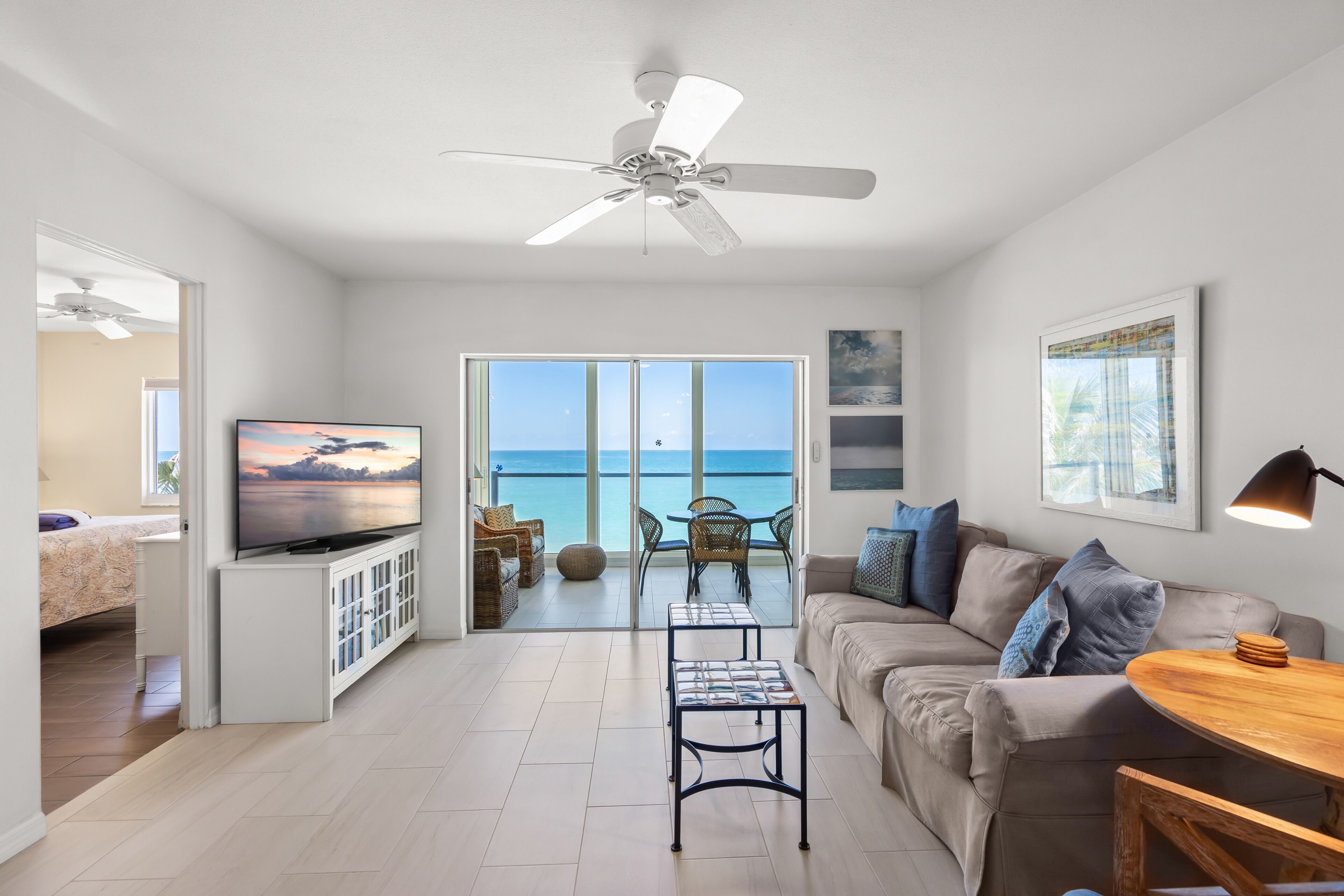 A modern living room with a television, a ceiling fan, a sofa, a coffee table, and a view of the ocean through large glass doors.