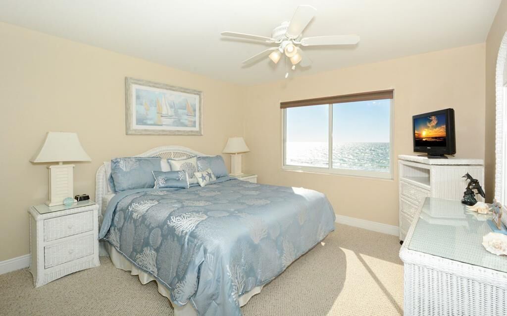 A bedroom with a bed, two bedside tables, a television, a window with a view, and a ceiling fan.