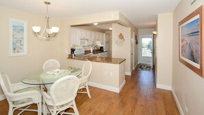 A dining area with a glass table and white wicker chairs, a kitchen with white cabinets, and a hallway leading to a door. - Gulf Front, 3rd Floor, 1 bdr - A307, Gorgeous View! (Siesta Key)