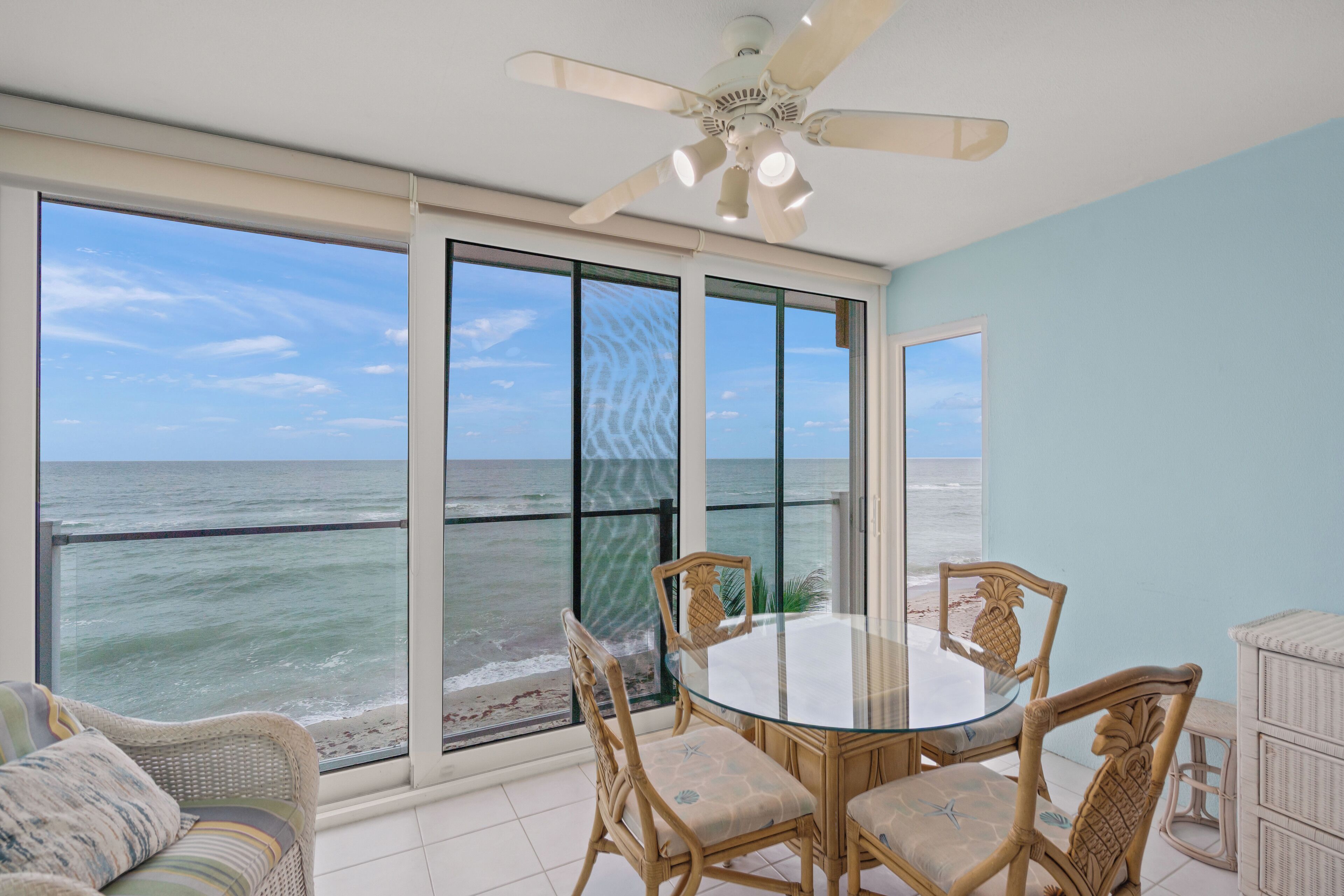 A room with a glass table and chairs, a ceiling fan, and a view of the ocean.