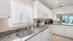 A modern kitchen with white cabinets, a granite countertop, stainless steel sink, and a view of the ocean through large windows. - Watch the Sunset from Your Gulf-Front Balcony (Siesta Key)