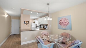 A dining area with wicker chairs and a glass table, a kitchen in the background, and a ceiling fan. - Watch the Sunset from Your Gulf-Front Balcony (Siesta Key)