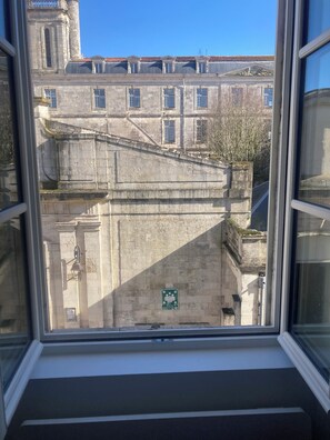 Blick durch ein Fenster eines Gebäudes mit einer steinernen Fassade, einem grünen Schild mit Symbolen und blauem Himmel.