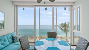 Una habitación con vista al mar, una mesa redonda de vidrio, sillas de mimbre y un sofá color turquesa.