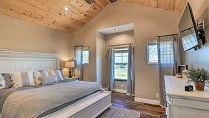 A bedroom with a large bed, wooden ceiling beams, a TV mounted on the wall, and a view through the door. - Pool | Hot Tub | Pickleball | Firepit (Williamstown)