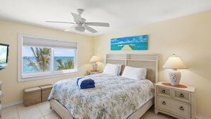 A bedroom with a bed, a ceiling fan, a window with a view of the ocean, two bedside lamps, and a painting on the wall. - Sun, Sand & Gulf-Front Views (Sarasota)