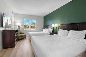 Ein Hotelzimmer mit zwei Betten, einem Schminktisch, einem Sessel, einem Schreibtisch mit Lampen und einem Fenster mit Jalousien.